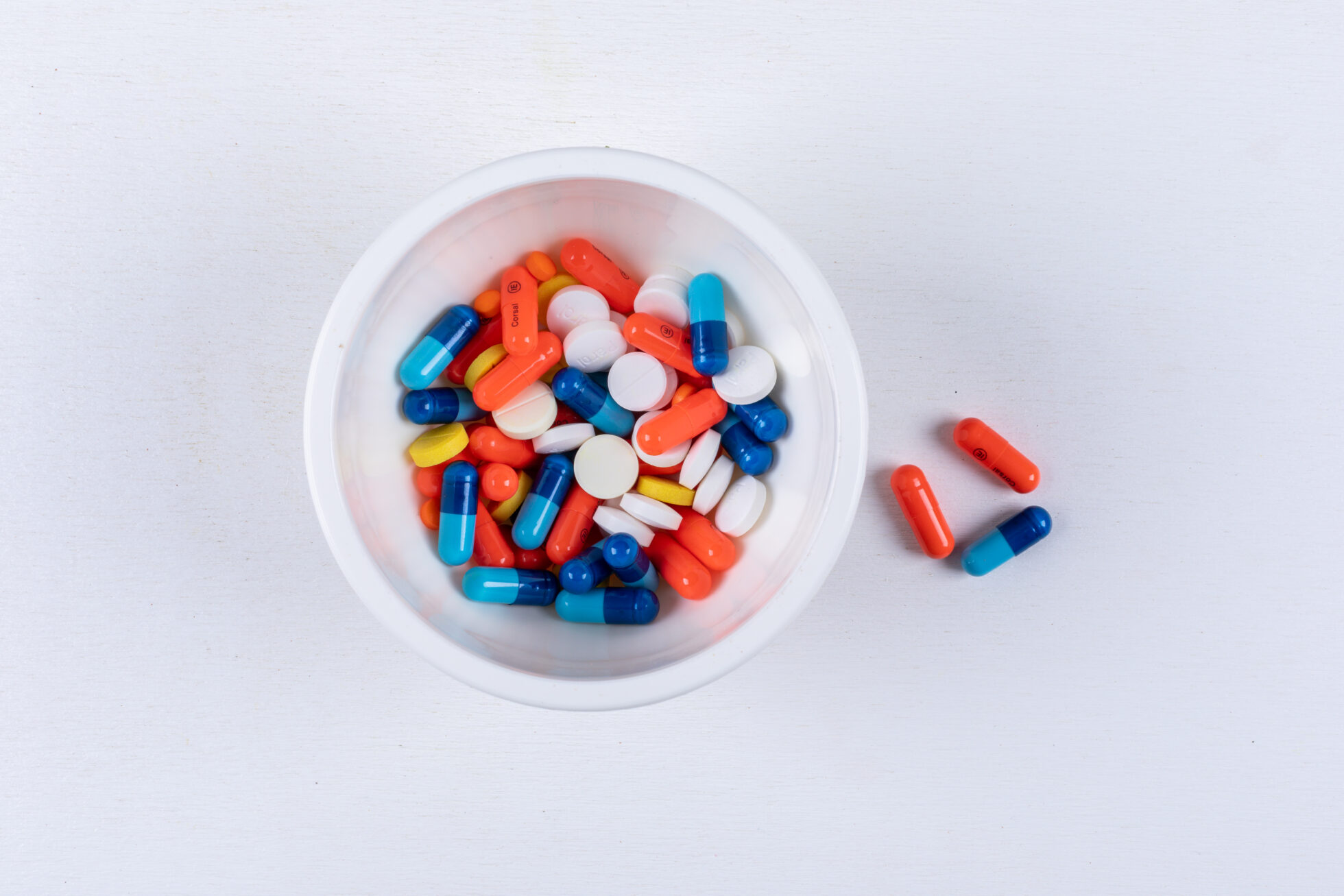 Top-down view of various pills and capsules in a bowl and scattered on a white surface
